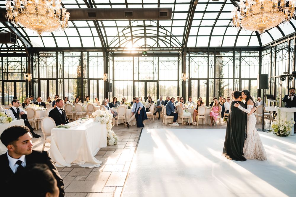Elegant wedding reception in a glass greenhouse venue with guests seated at white-draped tables as couple shares first dance.