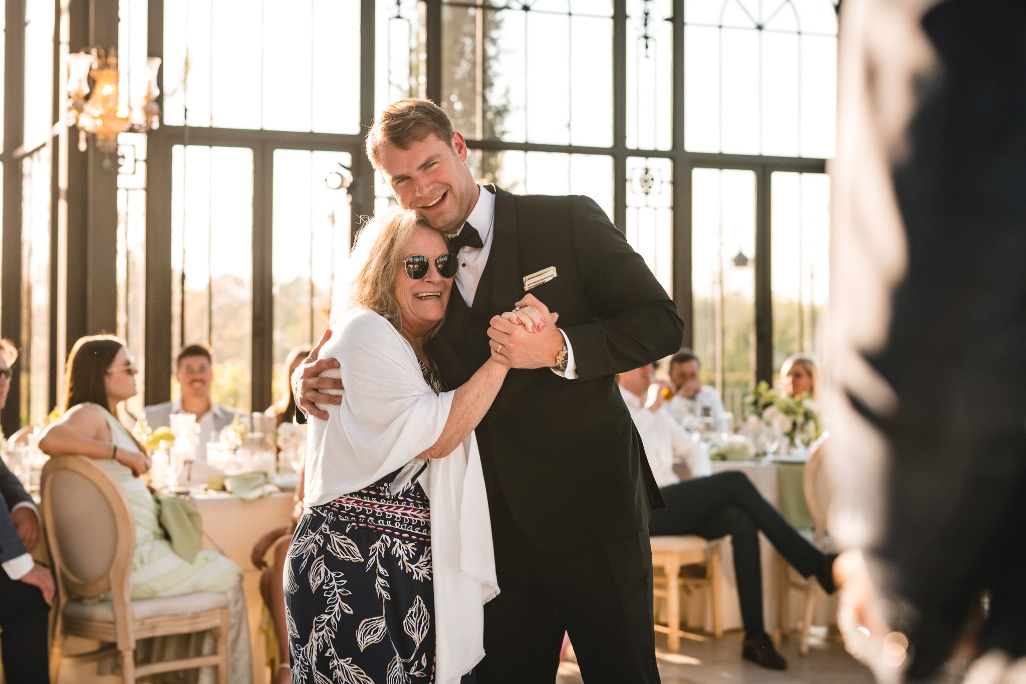 Groom shares a tender mother-son dance at elegant wedding reception with warm golden lighting.