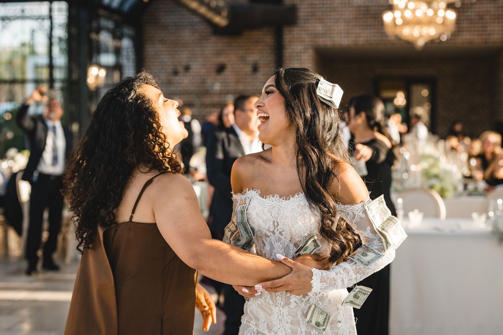 Bride laughs joyfully with guest holding dollar bills during lively wedding reception celebration.