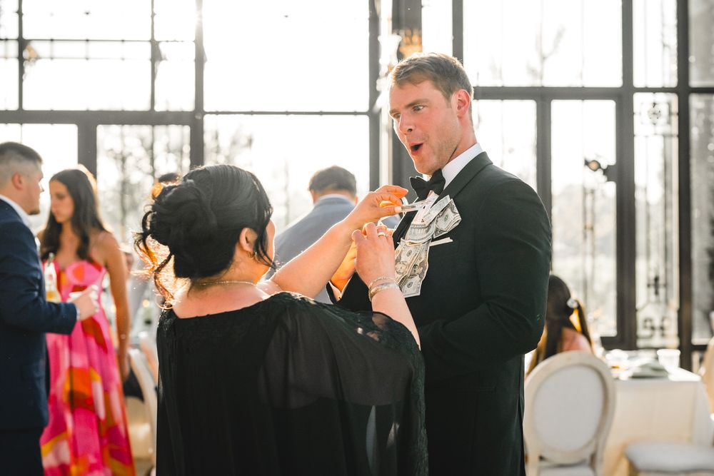 Groom in black tuxedo interacting with guests holding money at a lively wedding reception.