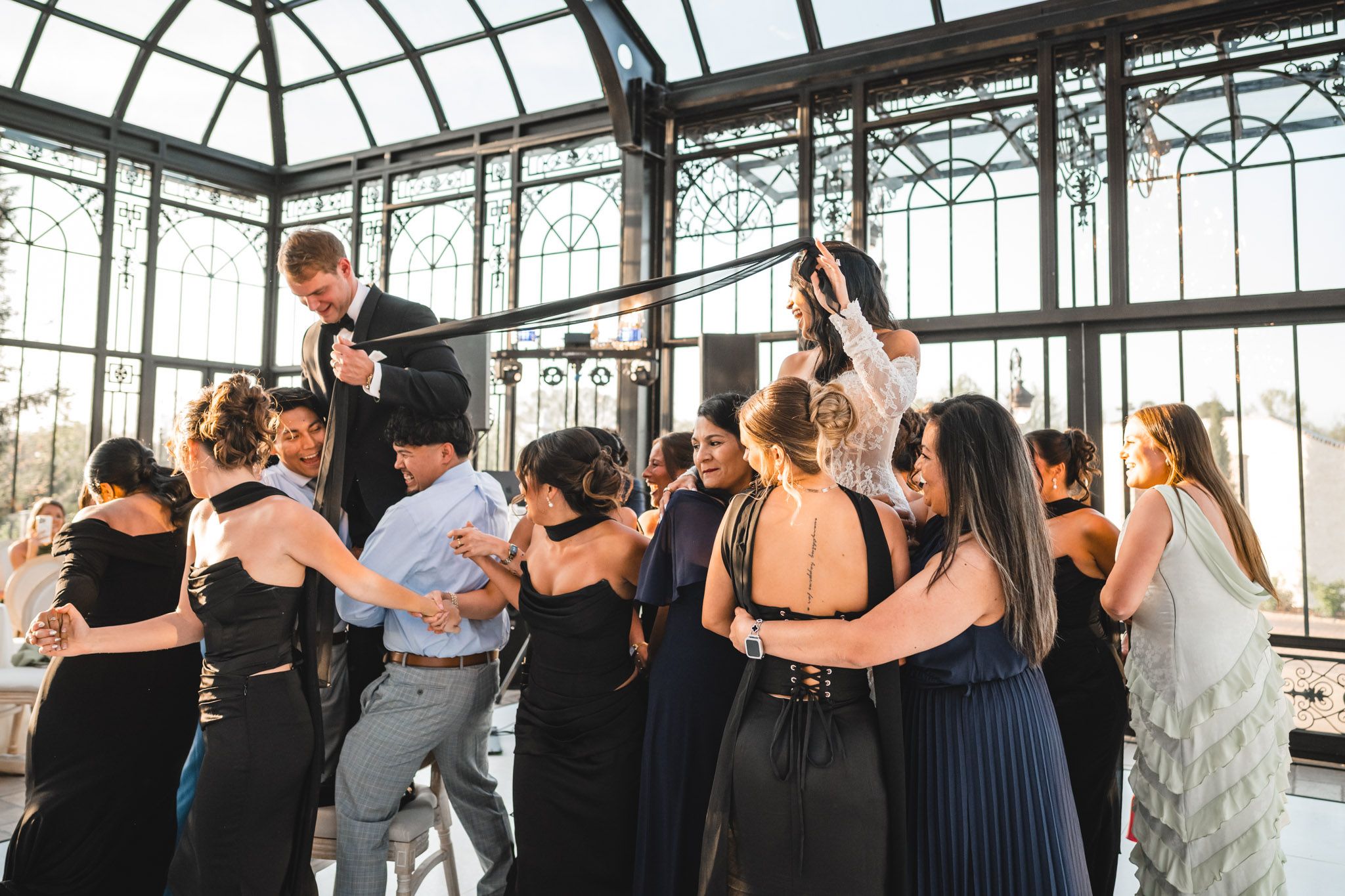 Energetic wedding guests dance together in a glass conservatory during lively reception celebration.