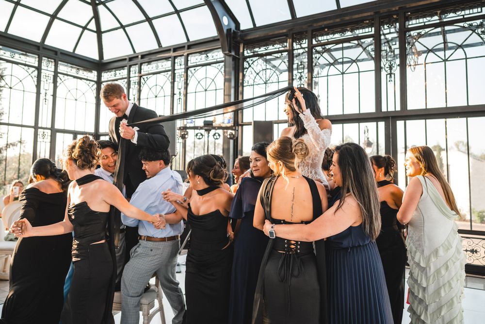 Energetic wedding guests dance together in a glass conservatory during lively reception celebration.