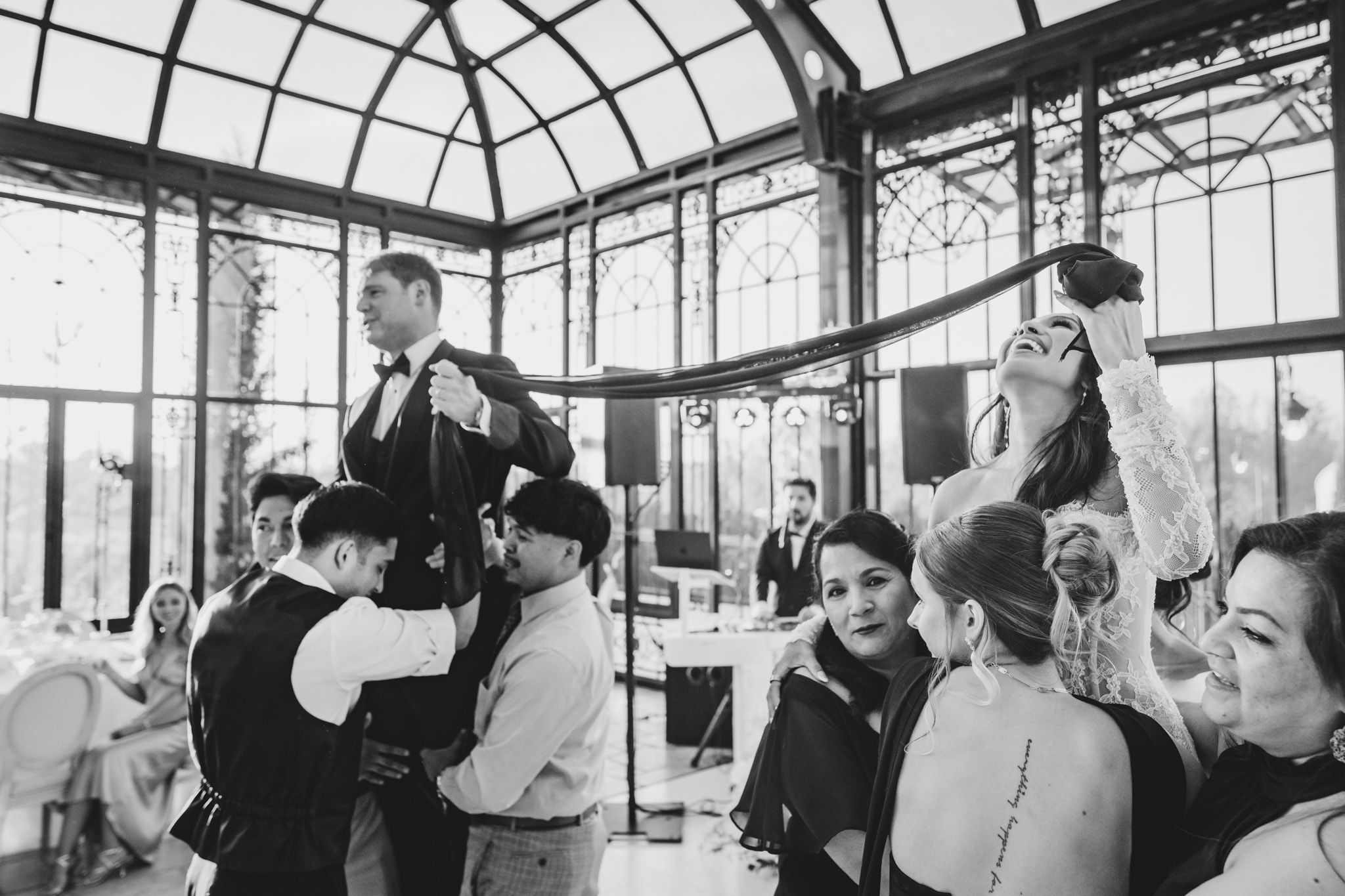 Black and white wedding photo of bride and groom being lifted by guests in elegant glass conservatory.