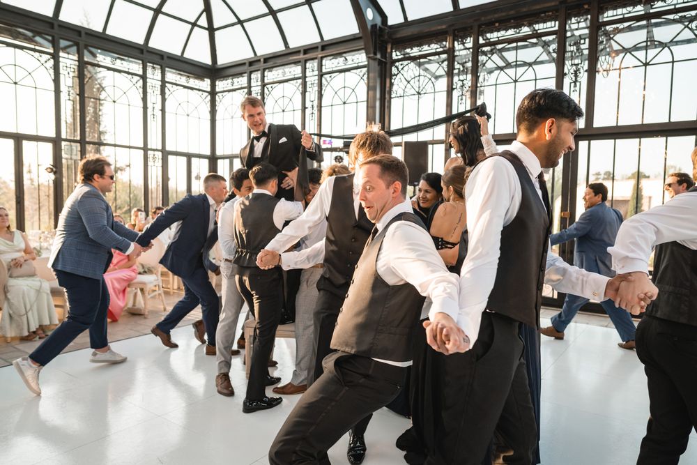 Groomsmen and guests perform lively line dance on white dance floor in glass greenhouse venue.