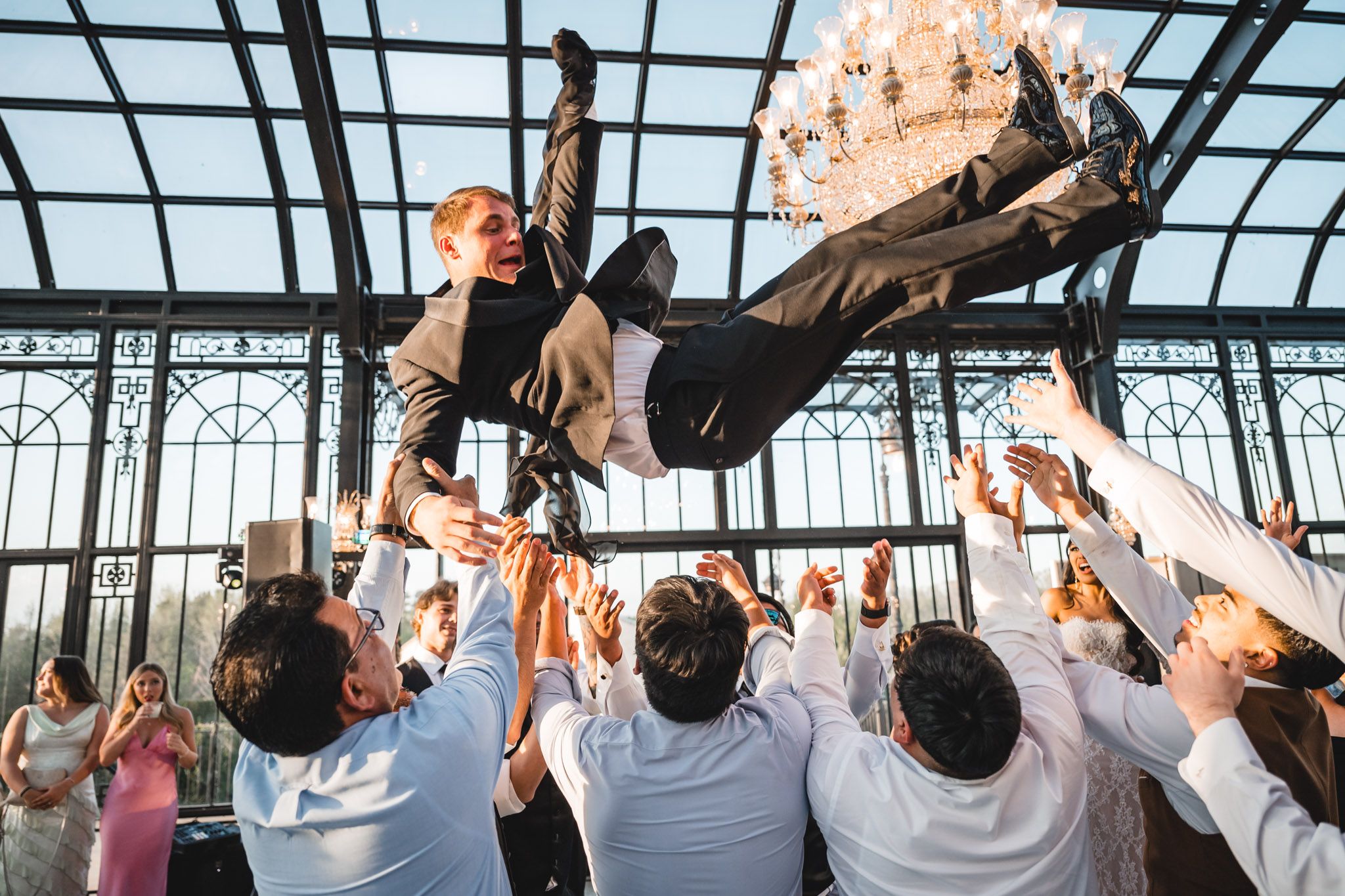 Groom being tossed in the air by groomsmen during a wedding reception in an elegant glass venue.