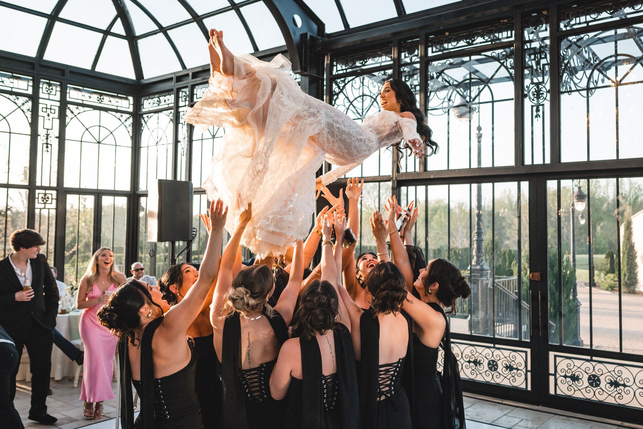 Bridesmaids in black dresses toss joyful bride into the air in stunning glass conservatory.