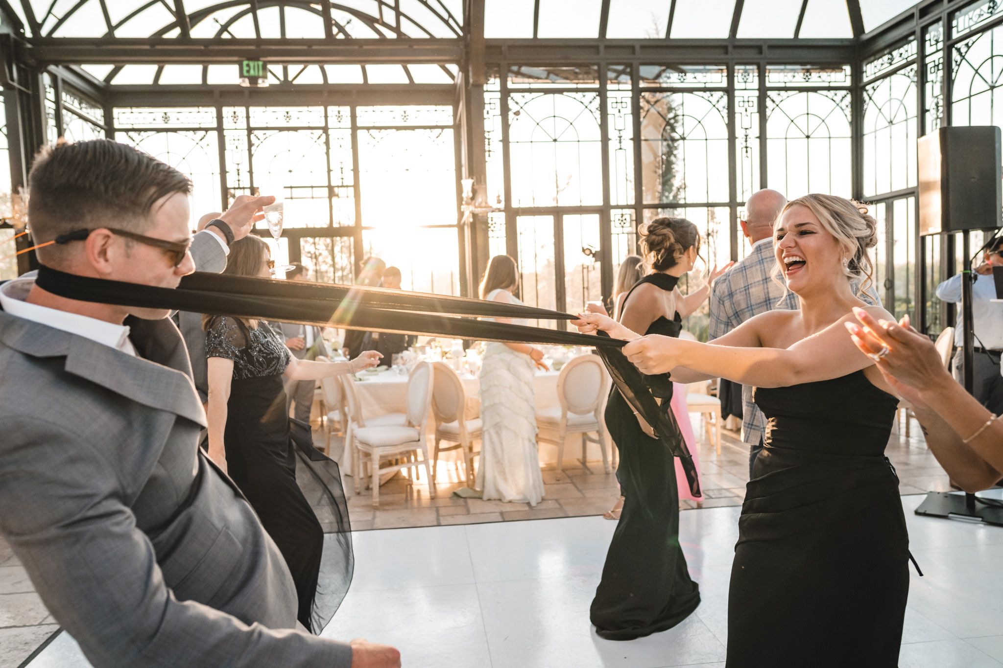 Guests laughing and dancing with a black ribbon on the dance floor of a sunlit greenhouse wedding venue.