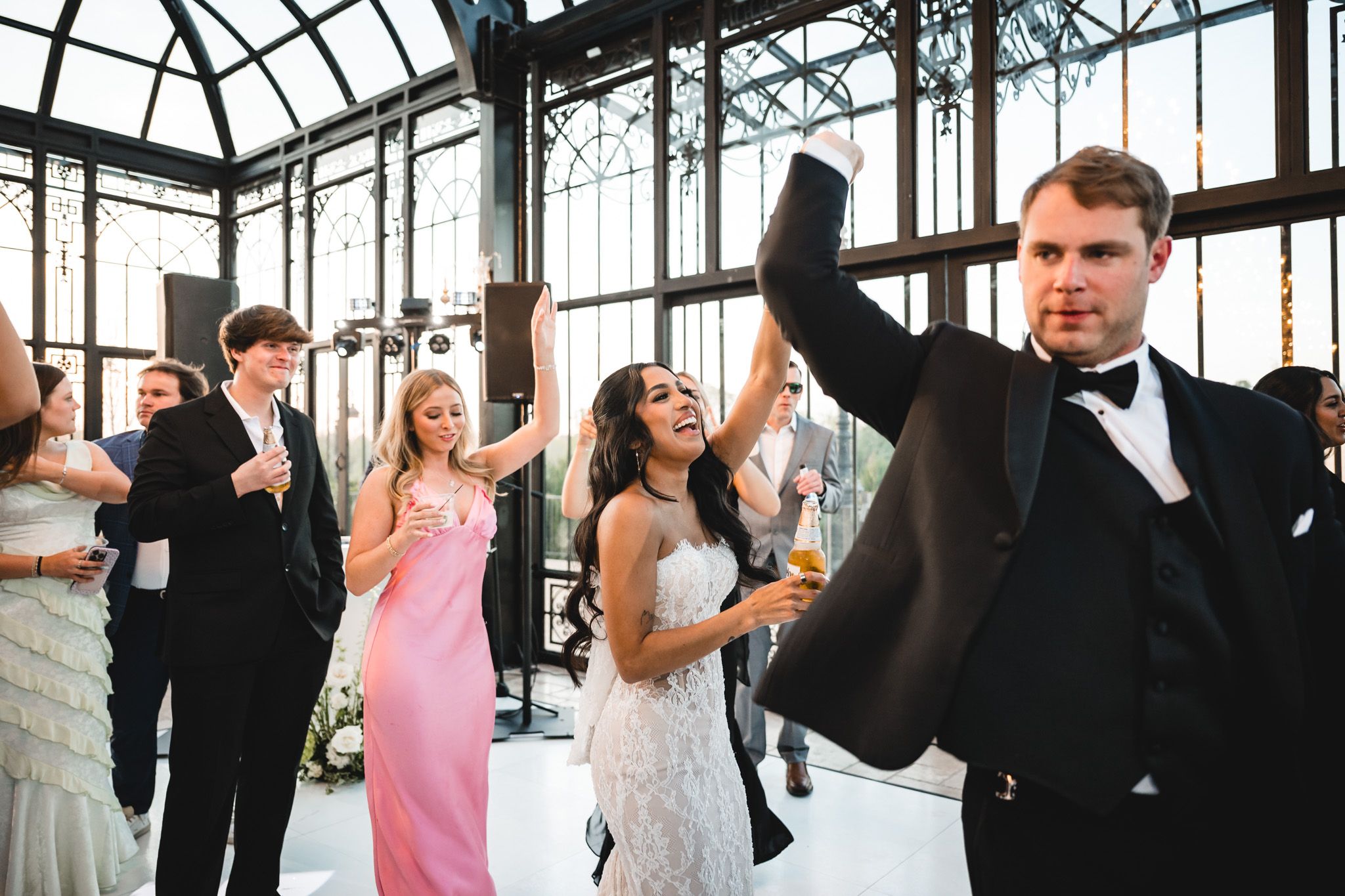 Wedding guests celebrate and dance in stunning glass conservatory venue during festive reception.