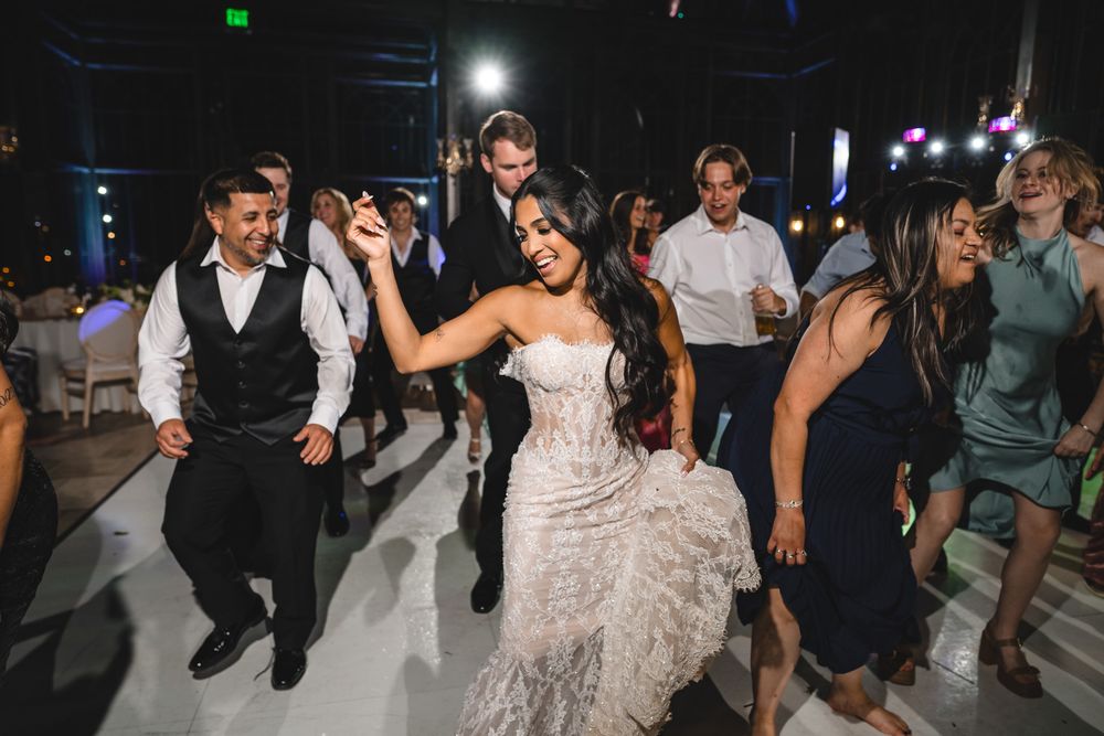 Bride in lace gown dances joyfully with guests on wedding reception dance floor.