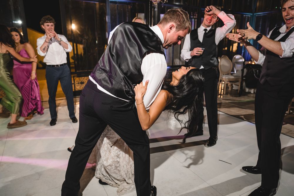 Groom dips bride dramatically on outdoor dance floor surrounded by cheering wedding guests at night.