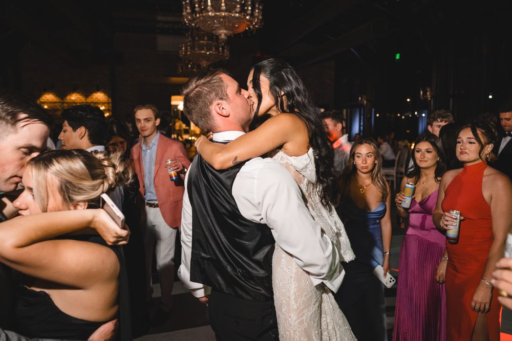 Bride and groom share romantic kiss surrounded by celebrating guests at evening wedding reception.