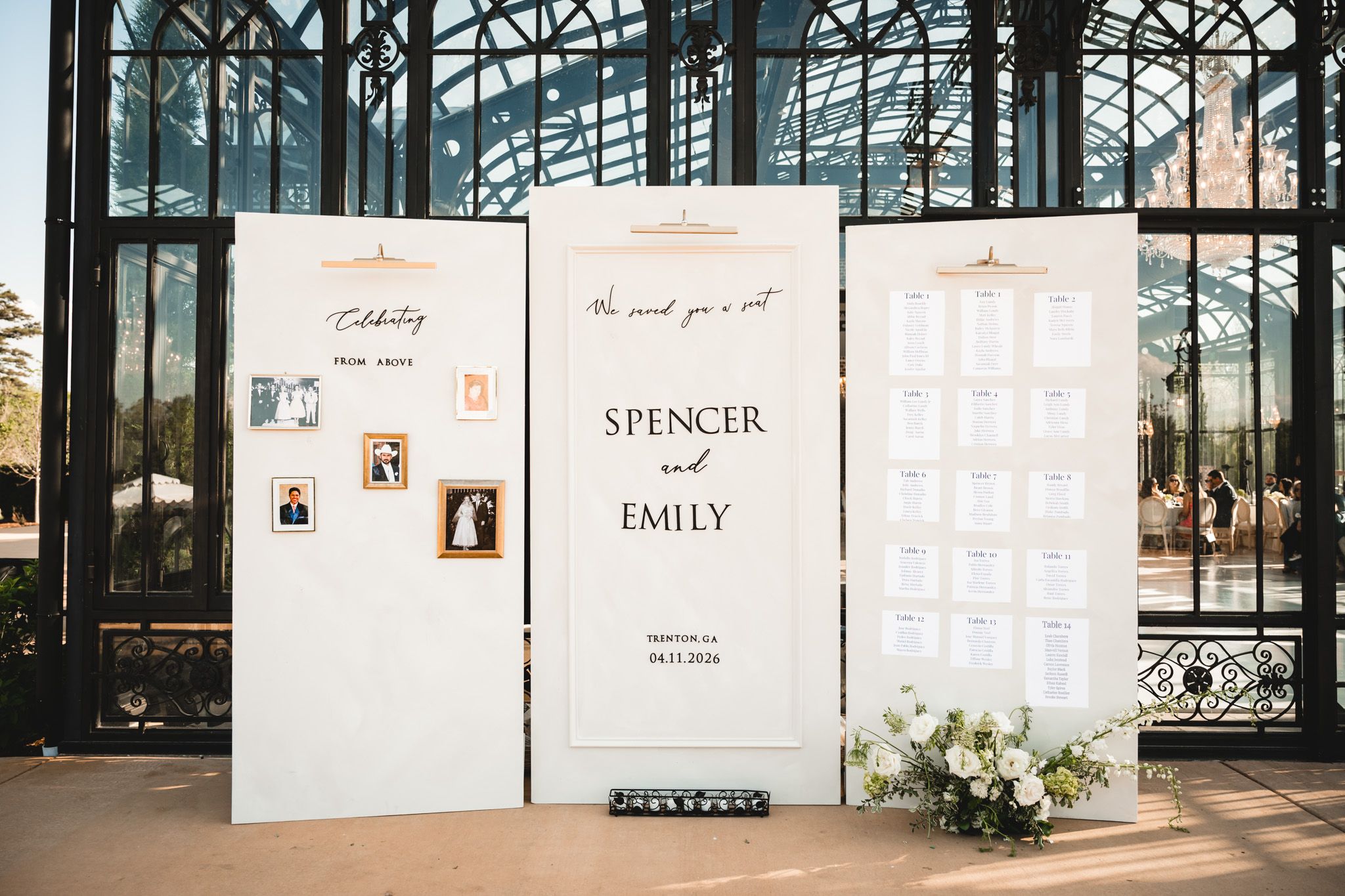 Elegant white tri-fold wedding seating chart display for Spencer and Emily with floral arrangements at a glass venue.