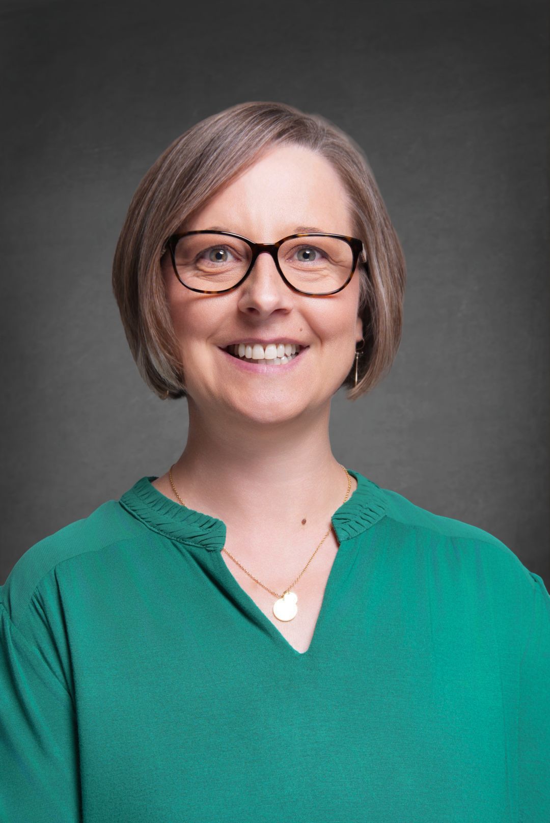 Smiling woman with a short bob haircut, dark-rimmed glasses and a green blouse wearing a pendant necklace against a gray backdrop.