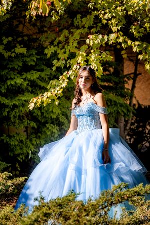 Young woman in flowing light blue quinceañera ball gown standing outdoors surrounded by lush green foliage in sunlight.