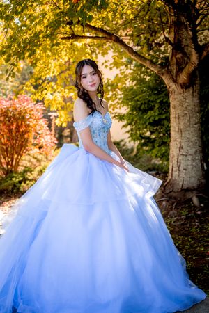 Young woman in a light blue off-shoulder ball gown poses outdoors under golden autumn trees.