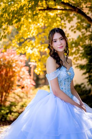 Teen girl in a blue beaded off-shoulder quinceañera gown smiles amid vibrant fall foliage.