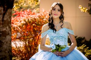 Young woman in light blue quinceañera ball gown holding white roses bouquet, posing outdoors with autumn foliage backdrop.