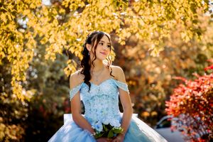 Young woman in light blue off-shoulder ball gown holding white bouquet, posing outdoors among golden autumn trees.