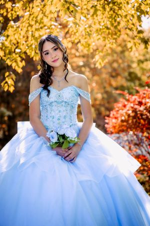 Young woman in a light blue quinceañera ball gown holding white roses, posing outdoors among autumn foliage.