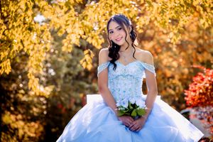 Smiling young woman in a blue quinceañera ball gown holds white roses outdoors in autumn.