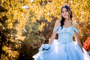 Young woman in a light blue off-shoulder quinceañera ball gown holding a bouquet, posing outdoors among autumn foliage.