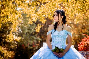 Young woman in elegant light blue off-shoulder ball gown holding bouquet among autumn foliage.
