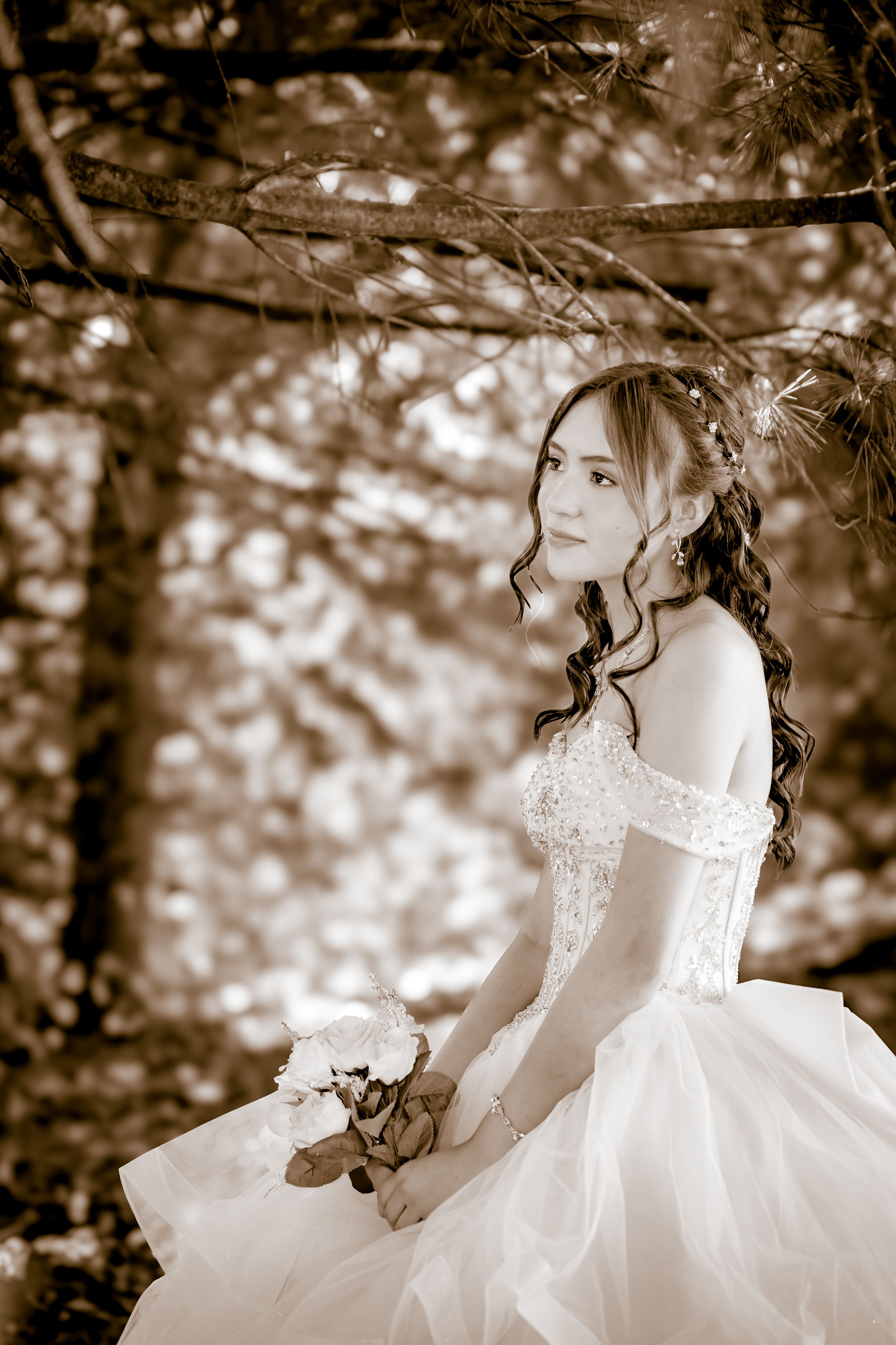 Sepia-toned portrait of a bride in an off-shoulder lace gown holding a white bouquet, posed under pine trees.