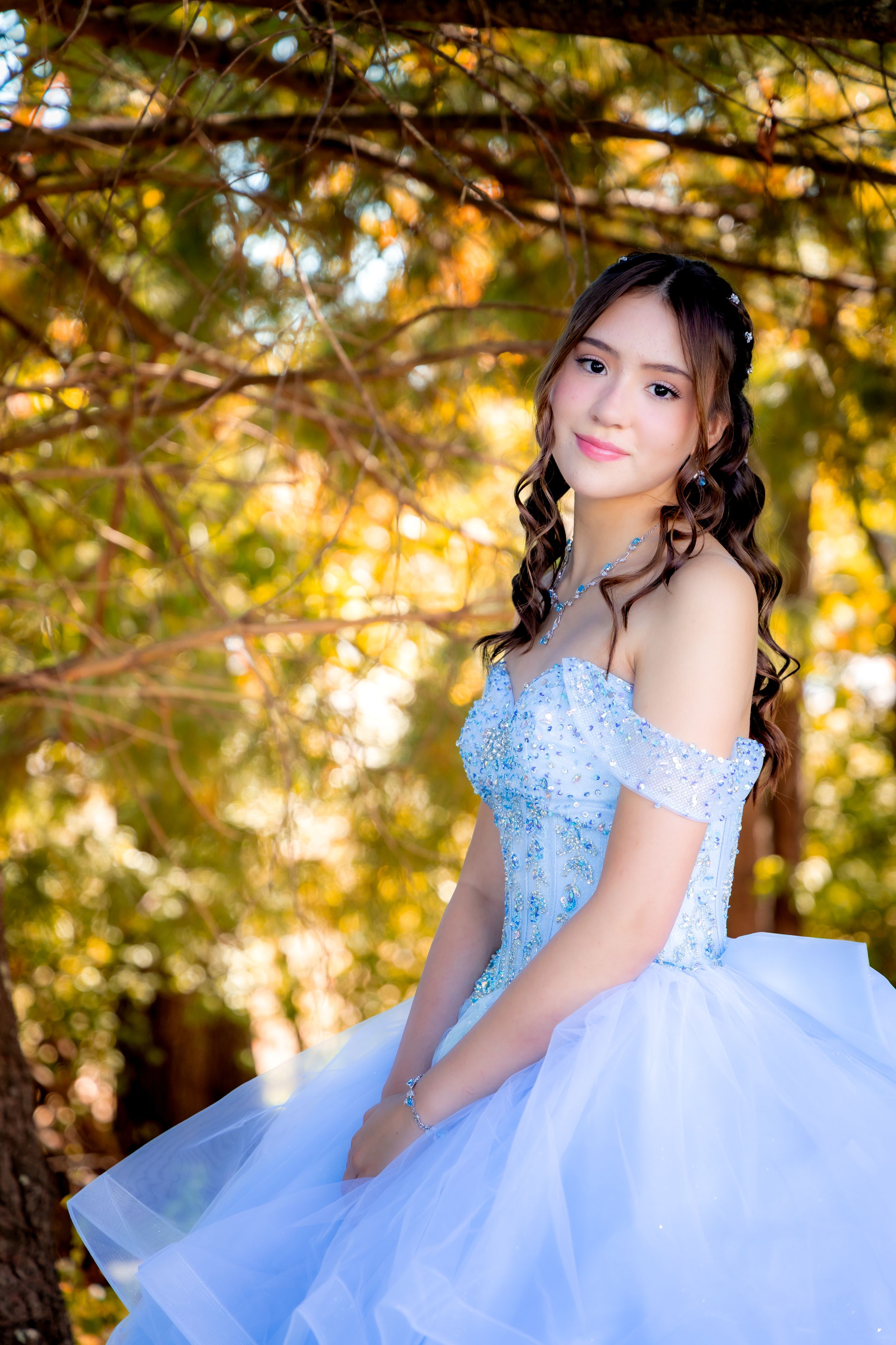 Young woman in light blue off-shoulder ball gown poses outdoors among autumn trees for quinceañera portrait.