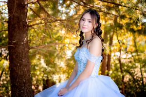 Young woman in a light blue quinceañera ball gown posing outdoors among autumn trees with golden foliage.