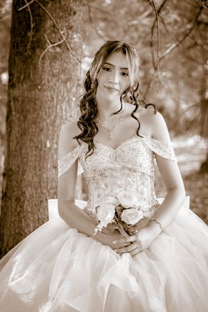 Sepia-toned portrait of a young woman in a beaded off-shoulder ball gown holding a white rose bouquet outdoors.