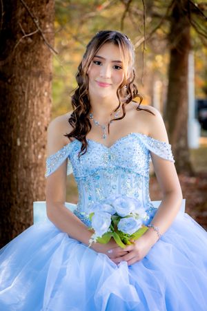 Young woman in light blue off-shoulder quinceañera ball gown holding white roses bouquet, seated outdoors among autumn trees.