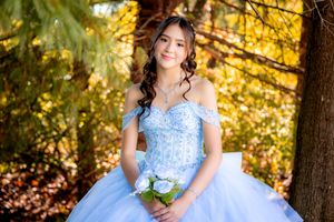 Young woman in a light blue ball gown quinceañera dress holds white roses bouquet outdoors in autumn forest setting.