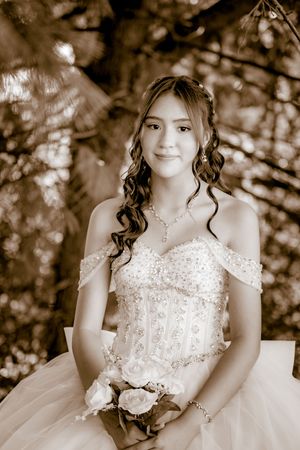 Young woman in beaded off-shoulder ball gown holding white roses, sepia-toned quinceañera portrait outdoors.