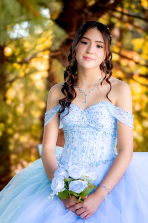 Teen girl in elegant light blue beaded quinceañera dress holding white rose bouquet with matching jewelry in autumn setting.