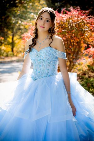 Young woman in a light blue off-shoulder ball gown poses outdoors among colorful autumn foliage.