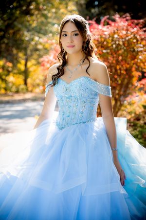 Young girl in a light blue off-shoulder ball gown with jeweled bodice poses outdoors among colorful autumn foliage.