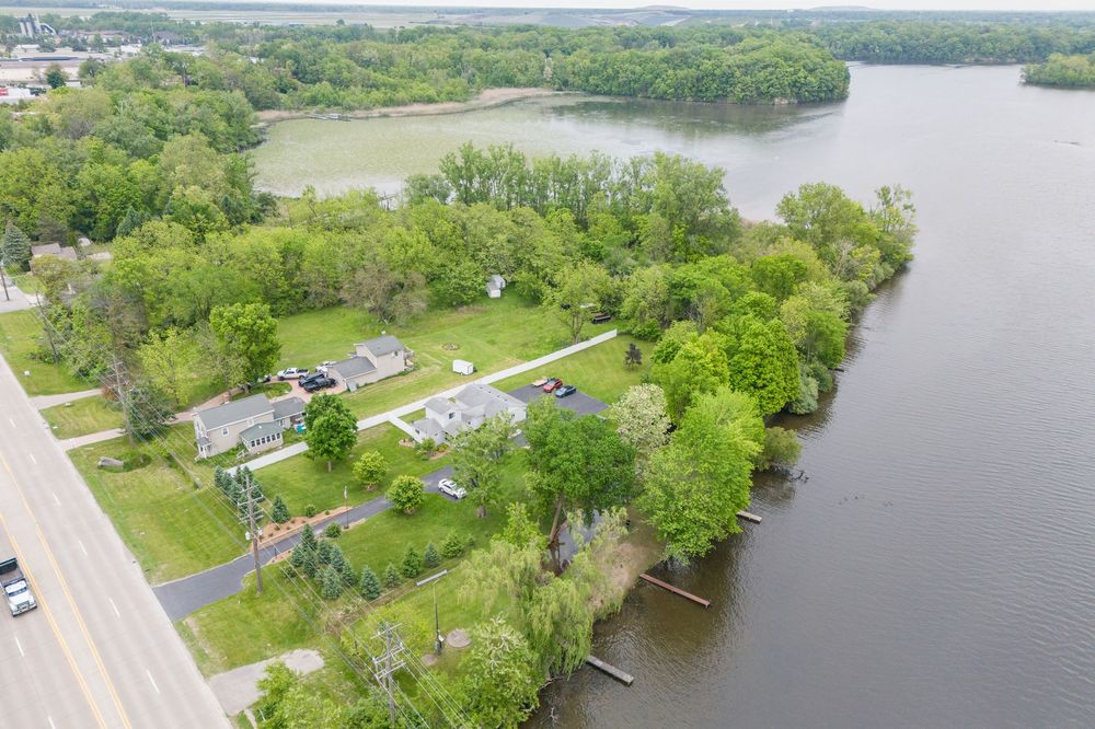 Aerial view of a waterfront neighborhood with tree-lined properties along a calm river and a paved road nearby.
