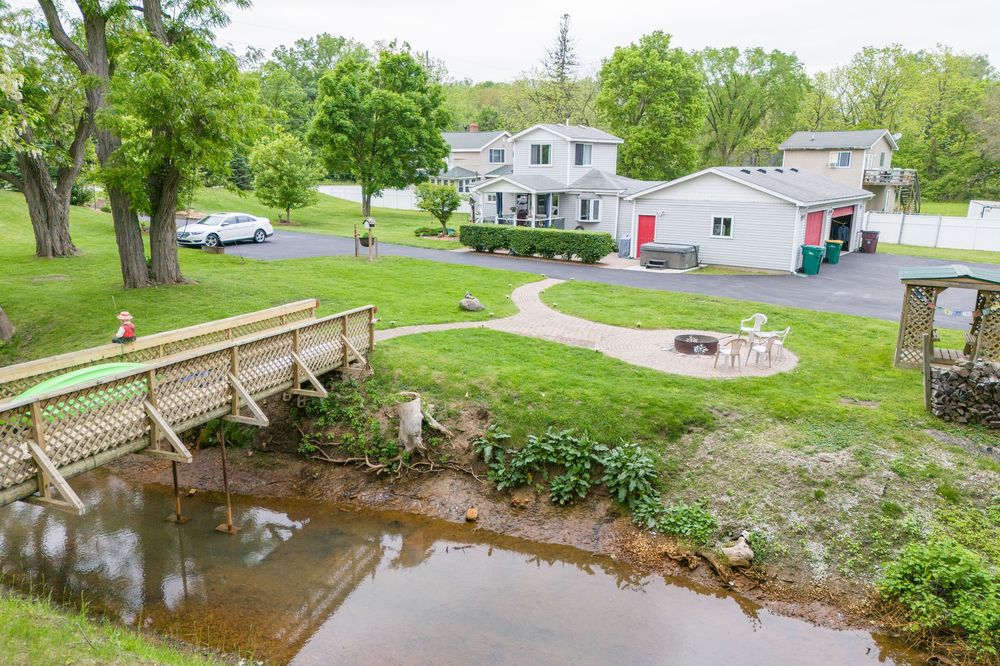 View of a two-story home with a wooden bridge over a small stream, patio area, and green lawn.
