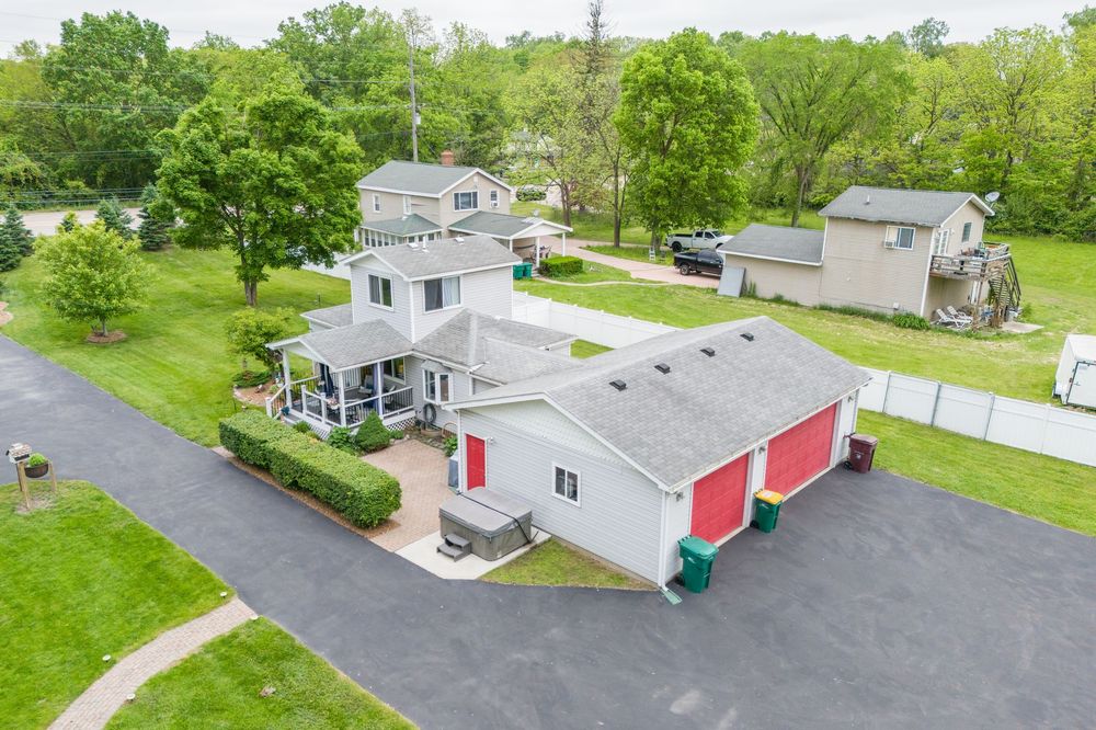 Aerial view of a two-story home with red garage doors, wraparound porch, and lush green trees in a suburban neighborhood.