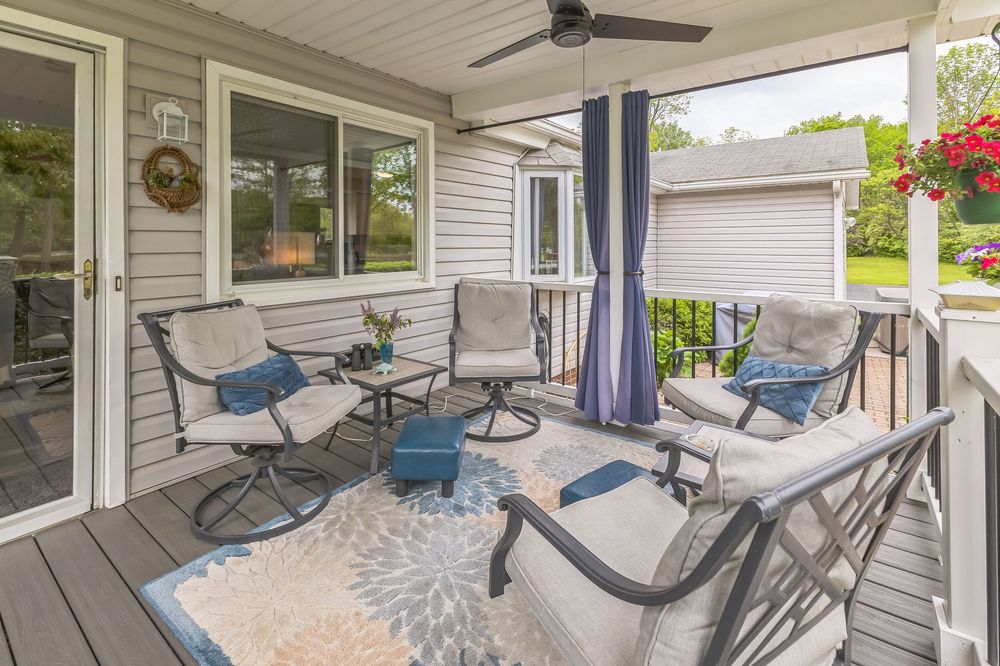 Spacious covered deck with cushioned patio furniture, ceiling fan, blue curtains, and lush garden views.
