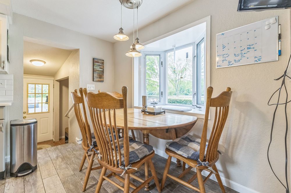 Bright dining area with round wooden table, spindle-back chairs, bay window, and pendant lights on hardwood floors.