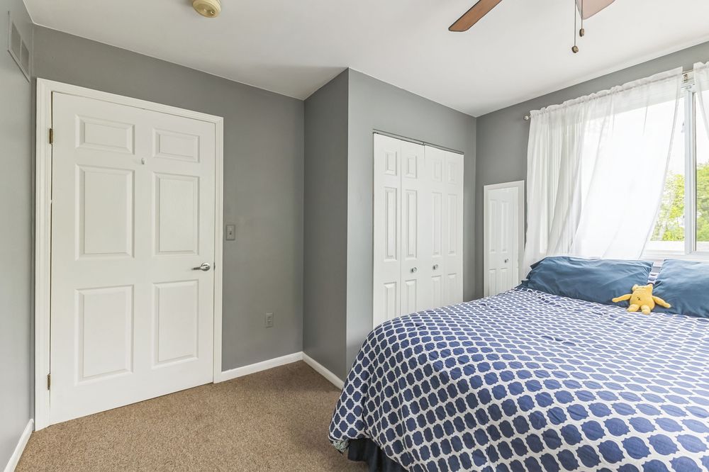 Cozy bedroom with gray walls, blue geometric bedspread, ceiling fan, carpet flooring, and two white panel doors.