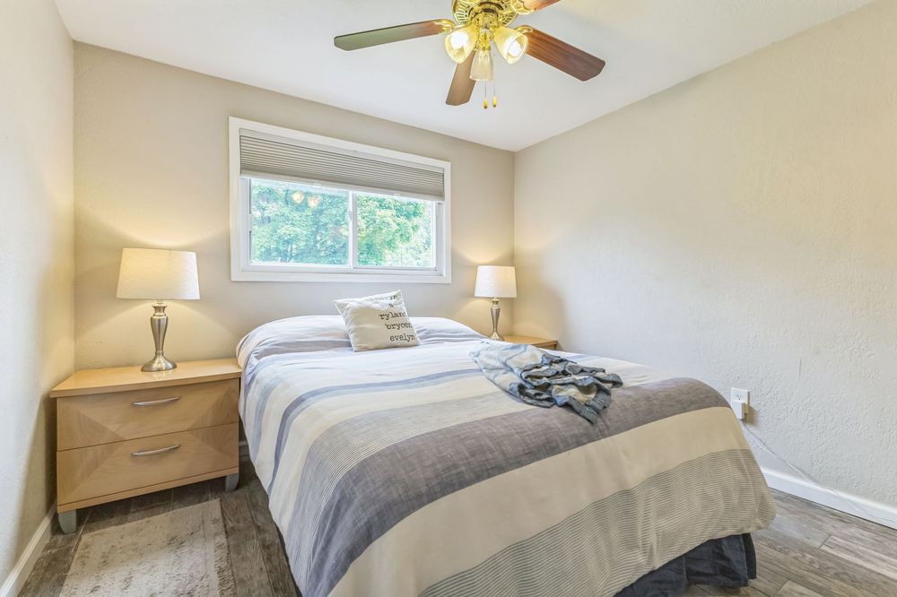 Bright bedroom with a queen bed, two nightstands with lamps, ceiling fan, and a window with natural light.