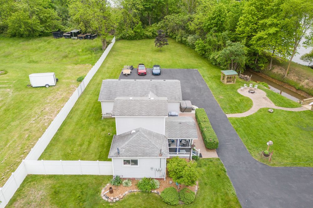 Aerial view of a suburban home with gray roof, black driveway, lush green lawn, gazebo, and surrounding trees.