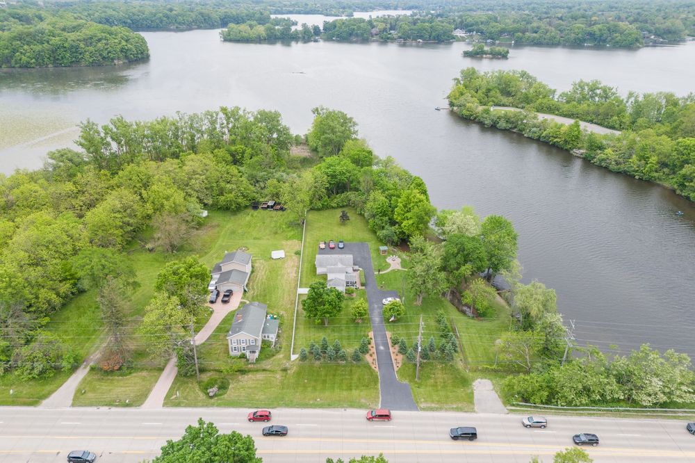 Aerial view of waterfront properties surrounded by lush trees along a river, with a road and passing cars visible below.