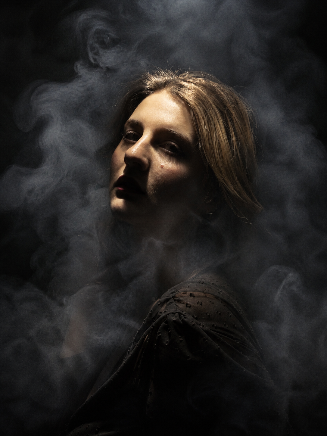 A woman emerges from swirling smoke in dramatic dark lighting, gazing upward with a mysterious, ethereal expression.