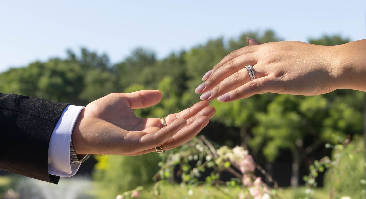 Close-up of two hands almost touching, with a diamond engagement ring on the woman's finger, outdoors.