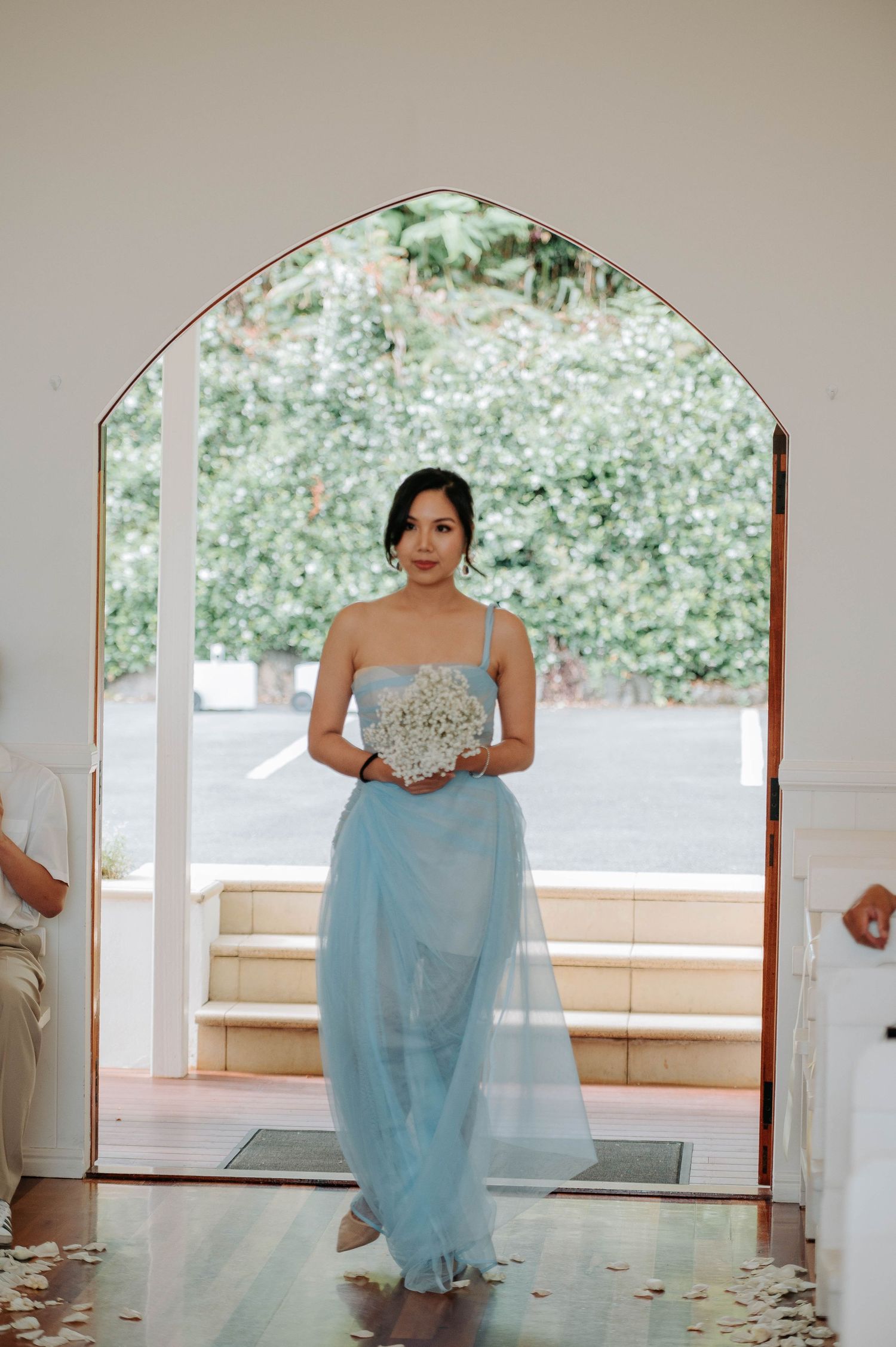 Bridesmaid in pale blue gown stands elegantly under arched doorway holding a bouquet.