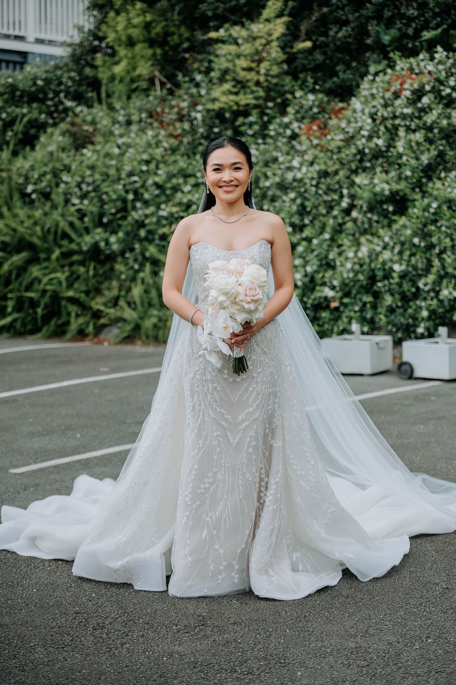 Bride in a stunning white lace gown with train poses outdoors against a garden backdrop.