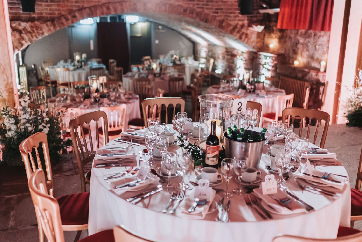 An elegant wedding reception venue with circular tables set with pink decor in a rustic brick cellar.
